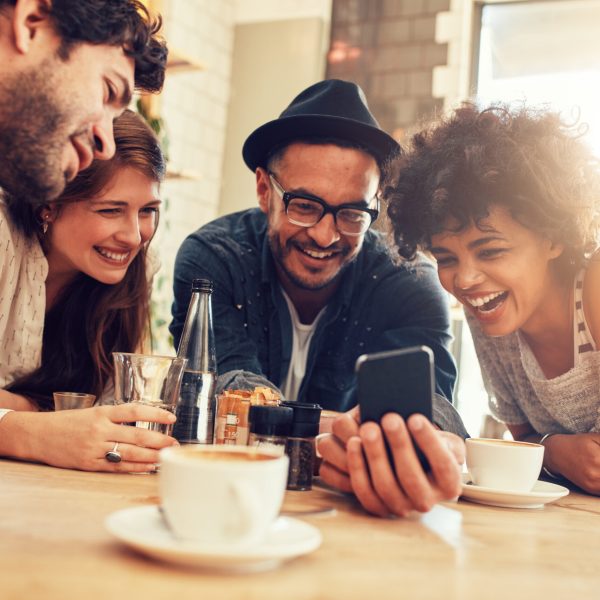 Portrait of cheerful young friends looking at smart phone while sitting in cafe. Mixed race people sitting at a table in restaurant using mobile phone.
